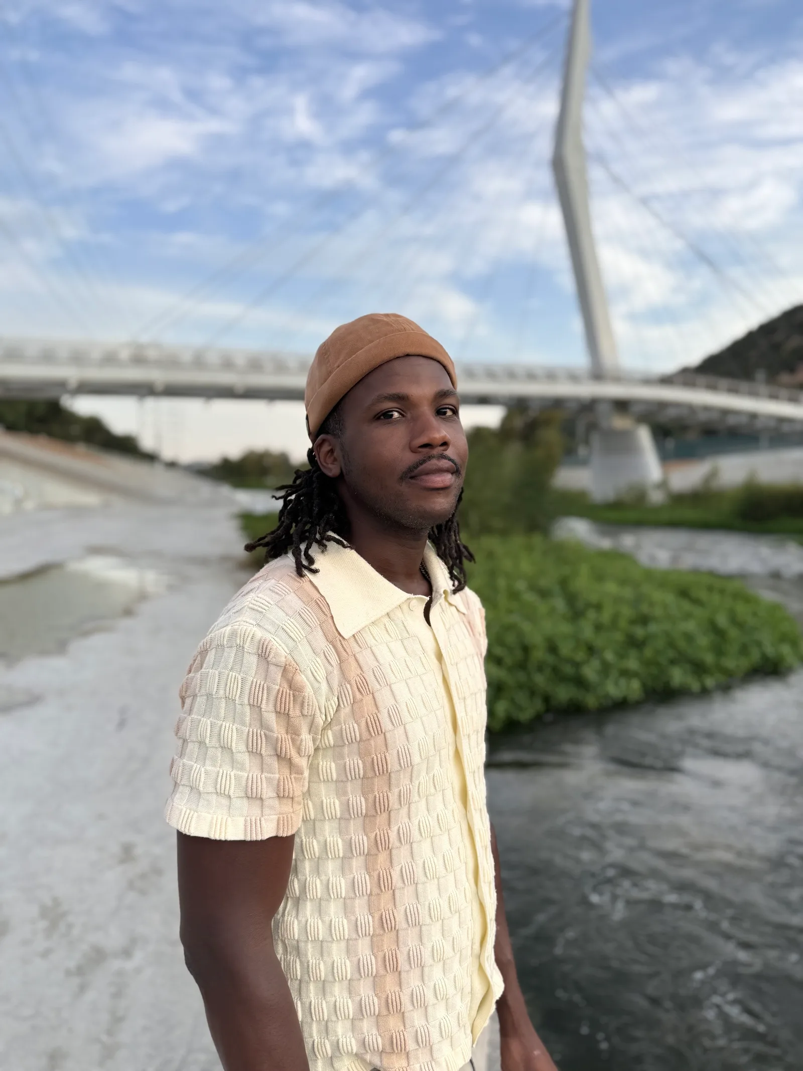 Bryan Brown, founder of R&B Consulting, wearing a cream shirt and brown cap beside a river and cable bridge