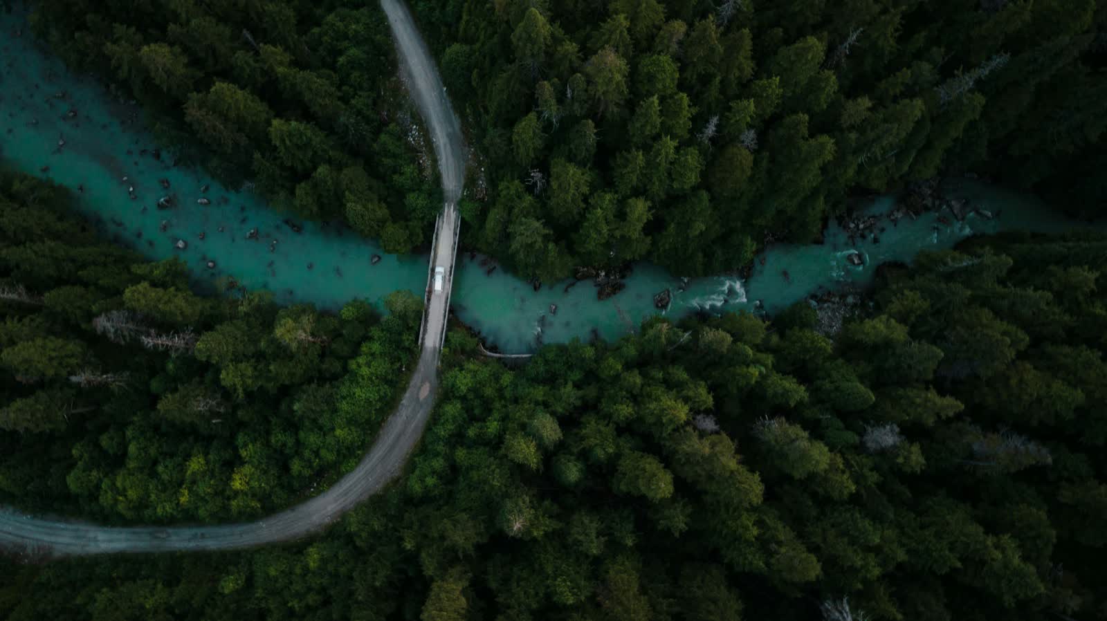 A bridge crossing a teal river winding through dark evergreen forest, seen from above